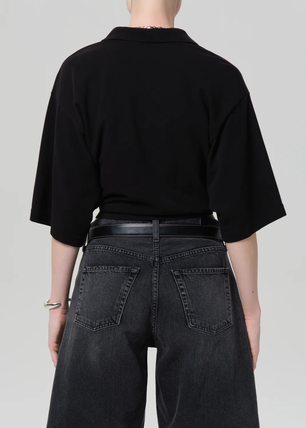 A person stands facing away, wearing a loose black lowen polo shirt by Citizens Of Humanity/AGOLDE with black jeans and a black belt against a plain light gray background.