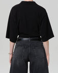 A person stands facing away, wearing a loose black lowen polo shirt by Citizens Of Humanity/AGOLDE with black jeans and a black belt against a plain light gray background.