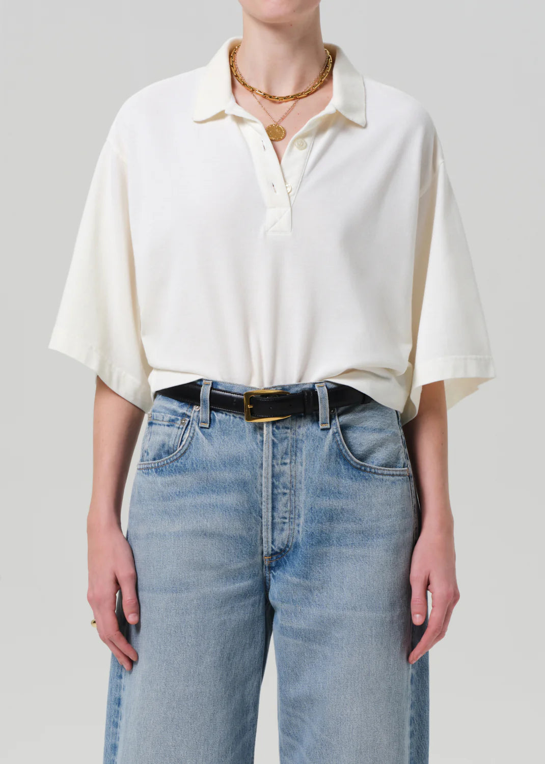 A person wears the Lowen Polo Shirt by Citizens Of Humanity/AGOLDE, tucked into high-waisted light blue jeans with a black belt, accessorized with layered gold necklaces and a ring, creating a feminine twist against a plain background.