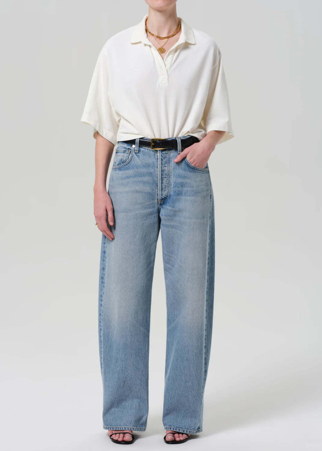 A person wears the Citizens Of Humanity/AGOLDE Lowen oversized white short-sleeve polo in breathable pique, paired with light blue high-waisted wide-leg jeans, a black belt, black heeled sandals, and layered gold necklaces against a plain light gray background.