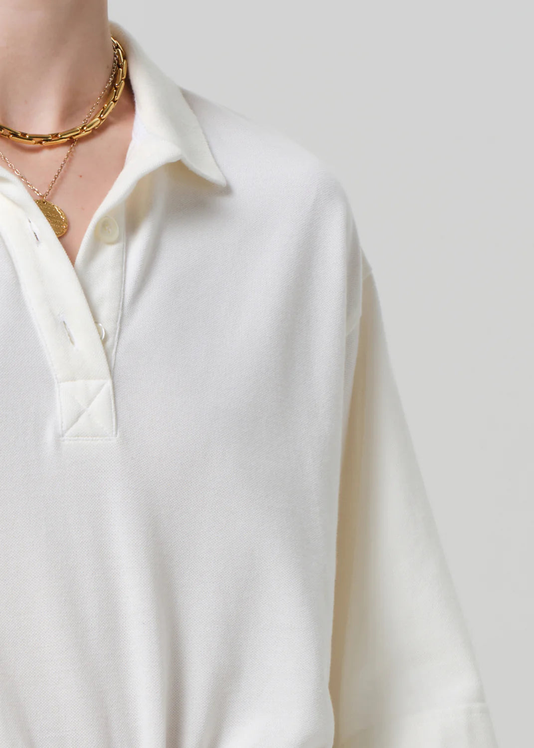 A person wears the cream Lowen Polo Shirt by Citizens Of Humanity/AGOLDE, featuring breathable pique fabric and buttons, styled with layered gold chain necklaces against a neutral backdrop; only their upper torso, neck, and shoulder are visible.