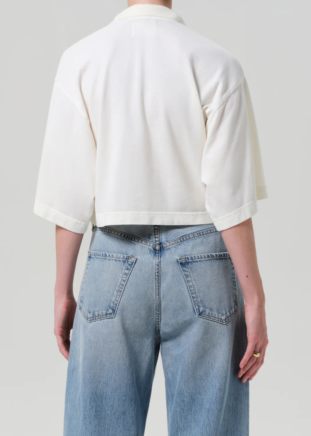 Someone is seen from the back against a plain light background, wearing Citizens Of Humanity/AGOLDE's Lowen Polo Shirt—a loose-fitting, cropped white pique top with elbow-length sleeves—paired with high-waisted, light blue jeans.