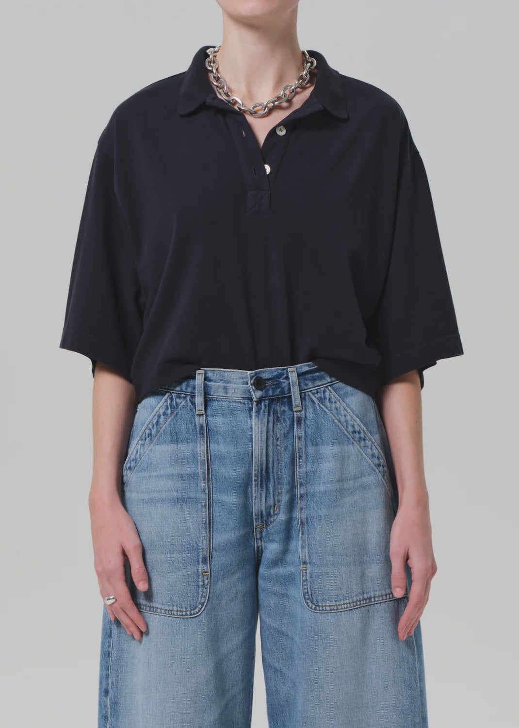 A person wears the Citizens Of Humanity/AGOLDE Lowen Polo Shirt, an oversized black pique top, tucked into high-waisted wide-leg blue jeans with front patch pockets, styled with a chunky silver chain necklace and ring against a plain light background.