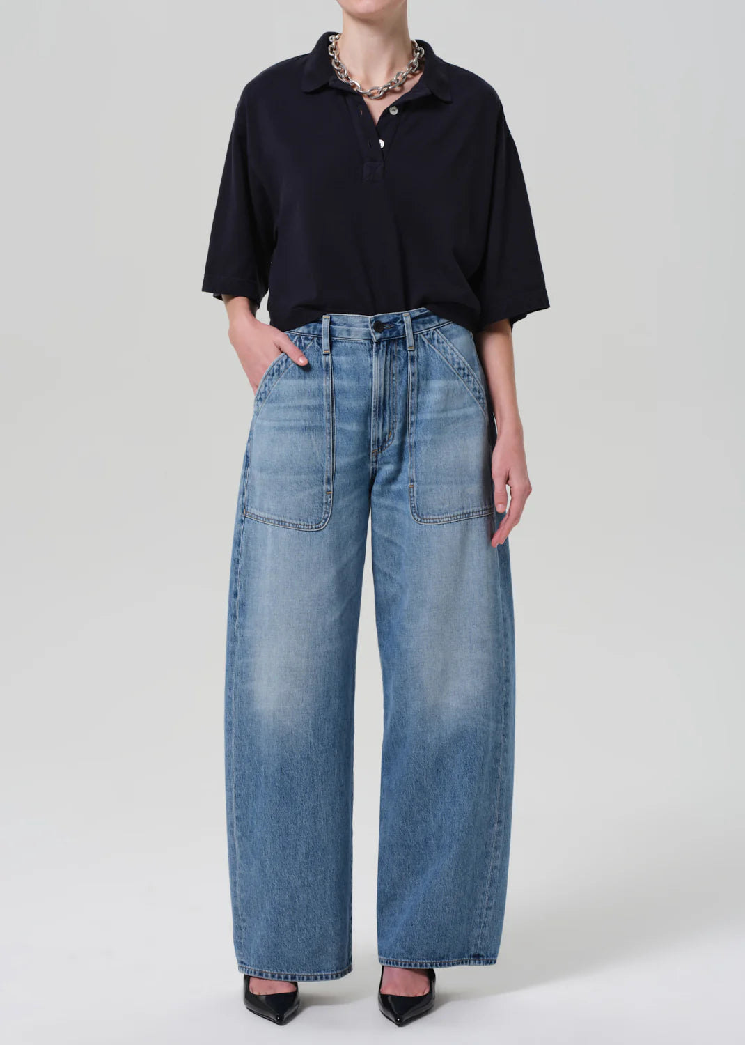 Model wears the Citizens Of Humanity/AGOLDE Lowen Polo Shirt—oversized, black, textured knit cotton—tucked into high-waisted, wide-leg blue jeans with large front pockets; paired with black pointed-toe heels against a plain light background.