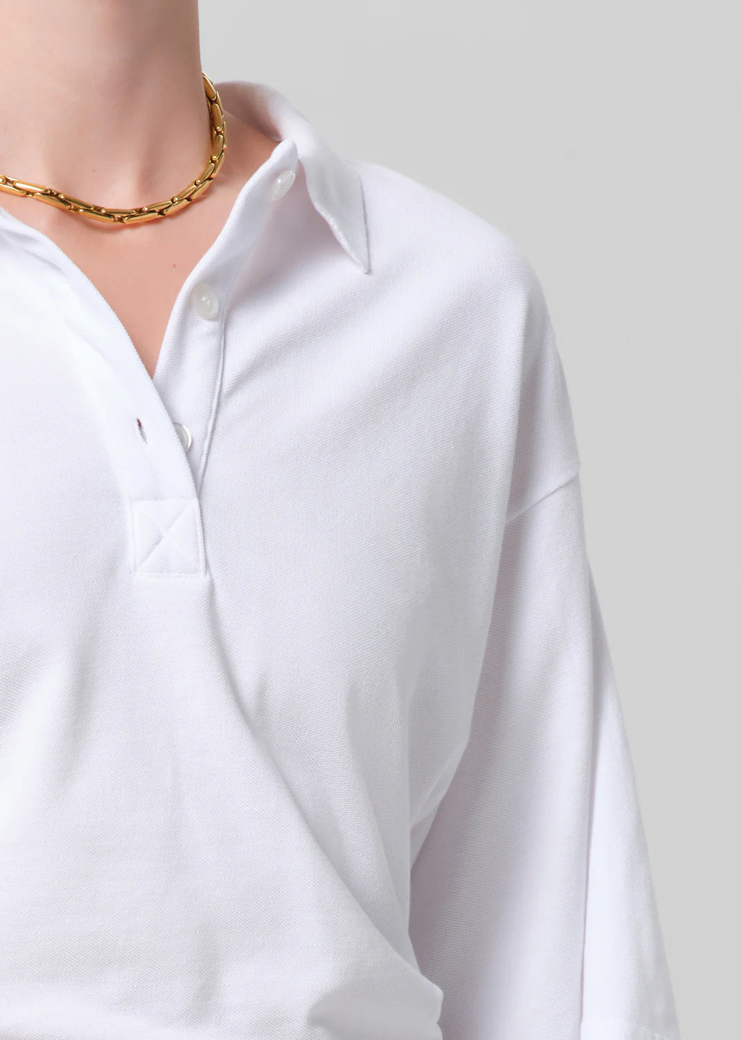 A person wears the Citizens Of Humanity/AGOLDE lowen polo shirt, a white short sleeve polo with buttons, paired with a gold chain necklace, shown from shoulders to mid-chest against a plain gray background.