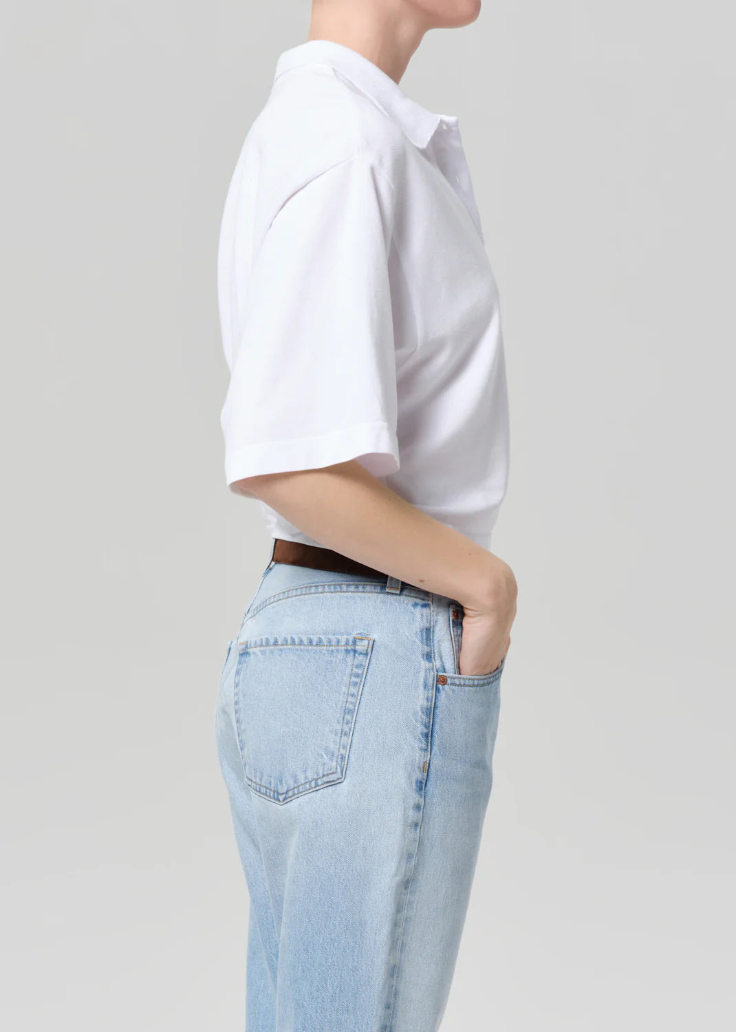 A person stands in profile with one hand in the pocket of light blue jeans, wearing a white Citizens Of Humanity/AGOLDE lowen polo shirt made from Supima jersey, set against a plain light gray background.