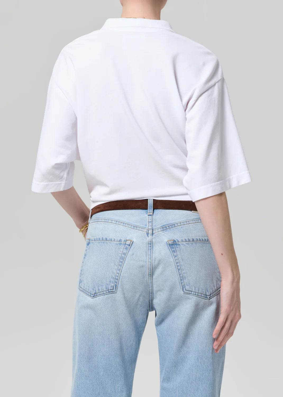 A person is seen from the back wearing a plain white Citizens Of Humanity/AGOLDE lowen polo shirt tucked into light blue high-waisted jeans with a brown belt, against a plain light gray background.