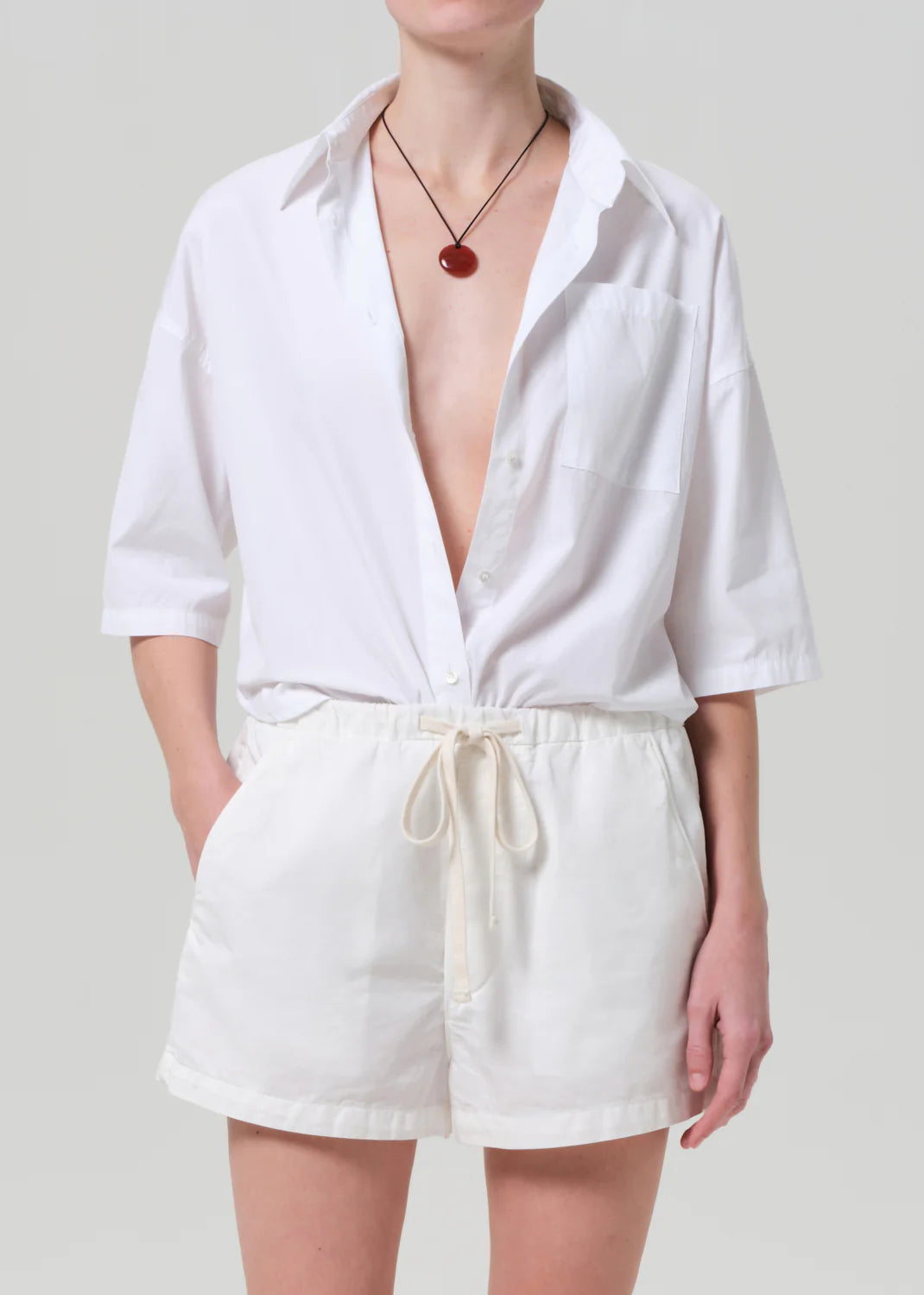 Wearing the Frances Cropped Cape Button Up by Citizens Of Humanity/AGOLDE, partially open and tucked into white drawstring shorts, with a red pendant necklace, a person stands before a plain light background.