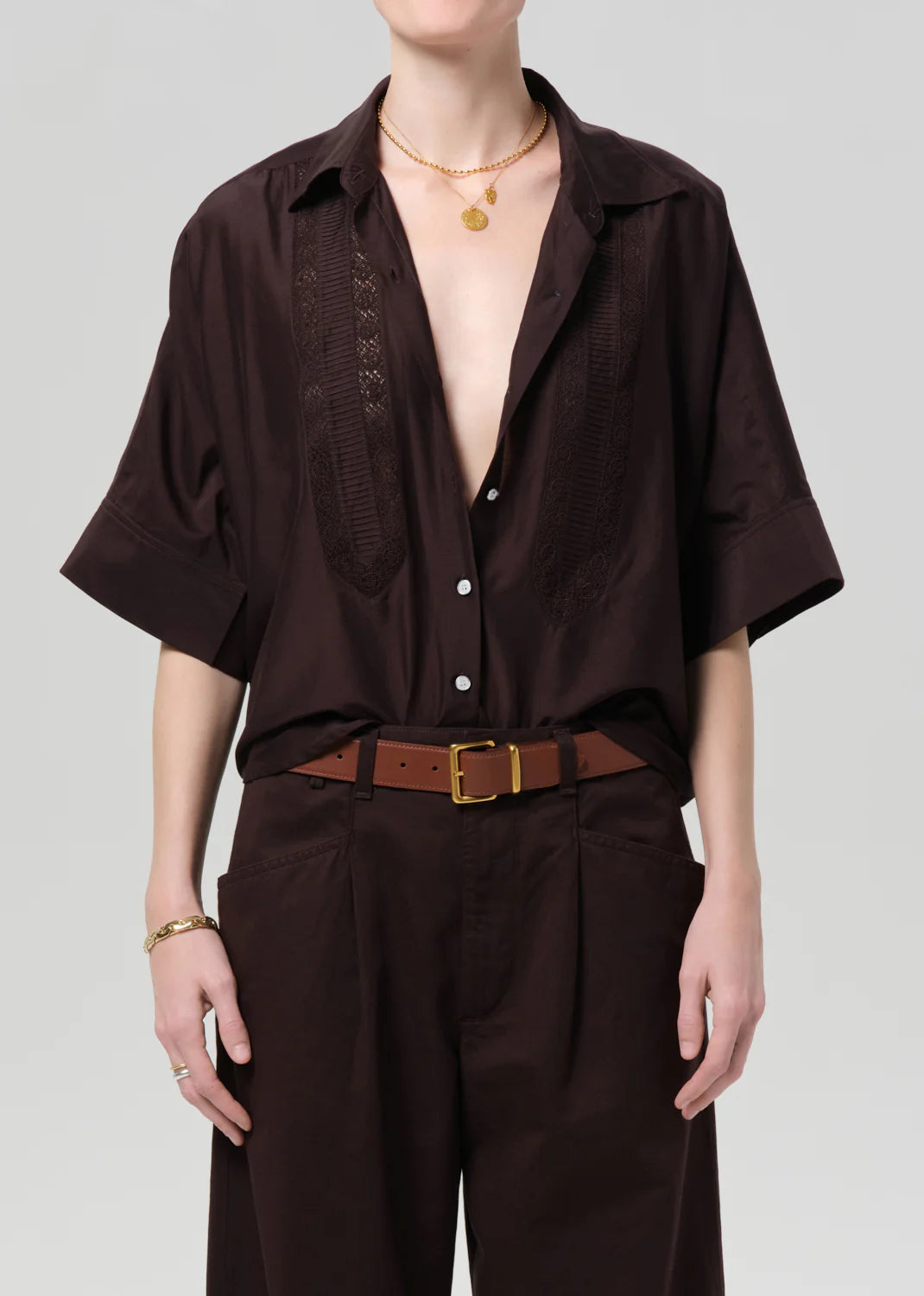 A person in a dark brown Claire with Lace Inset shirt by Citizens Of Humanity/AGOLDE, soft cotton silk blend and deep neckline, paired with brown high-waisted trousers, a brown belt with gold buckle, and layered gold necklaces on a light gray background.