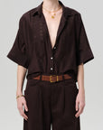 A person in a dark brown Claire with Lace Inset shirt by Citizens Of Humanity/AGOLDE, soft cotton silk blend and deep neckline, paired with brown high-waisted trousers, a brown belt with gold buckle, and layered gold necklaces on a light gray background.