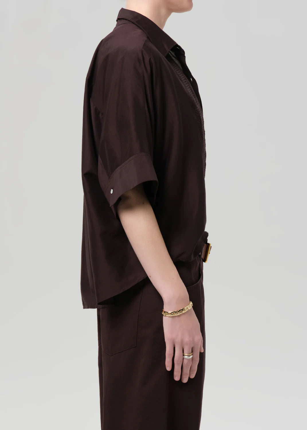 A person stands in profile against a plain background, wearing a loose-fitting, dark brown Claire with Lace Inset shirt and matching pants by Citizens Of Humanity/AGOLDE. They wear gold jewelry on their left hand, with their face partially out of frame.