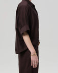 A person stands in profile against a plain background, wearing a loose-fitting, dark brown Claire with Lace Inset shirt and matching pants by Citizens Of Humanity/AGOLDE. They wear gold jewelry on their left hand, with their face partially out of frame.