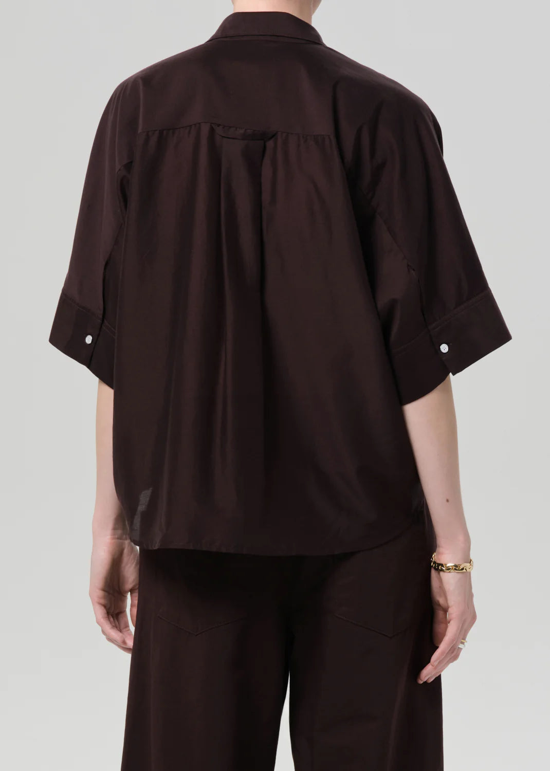 A person with short hair and a gold bracelet is seen from the back wearing Citizens Of Humanity/AGOLDE's Claire with Lace Inset—a loose, dark brown cotton-silk shirt featuring a yoke detail, lace inset, and white sleeve buttons—paired with matching pants.