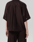 A person with short hair and a gold bracelet is seen from the back wearing Citizens Of Humanity/AGOLDE's Claire with Lace Inset—a loose, dark brown cotton-silk shirt featuring a yoke detail, lace inset, and white sleeve buttons—paired with matching pants.