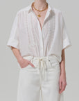 A person wears the Claire with Lace Inset by Citizens Of Humanity/AGOLDE, a loose short-sleeve shirt with lace details, tucked into high-waisted white pants and accessorized with layered gold necklaces.