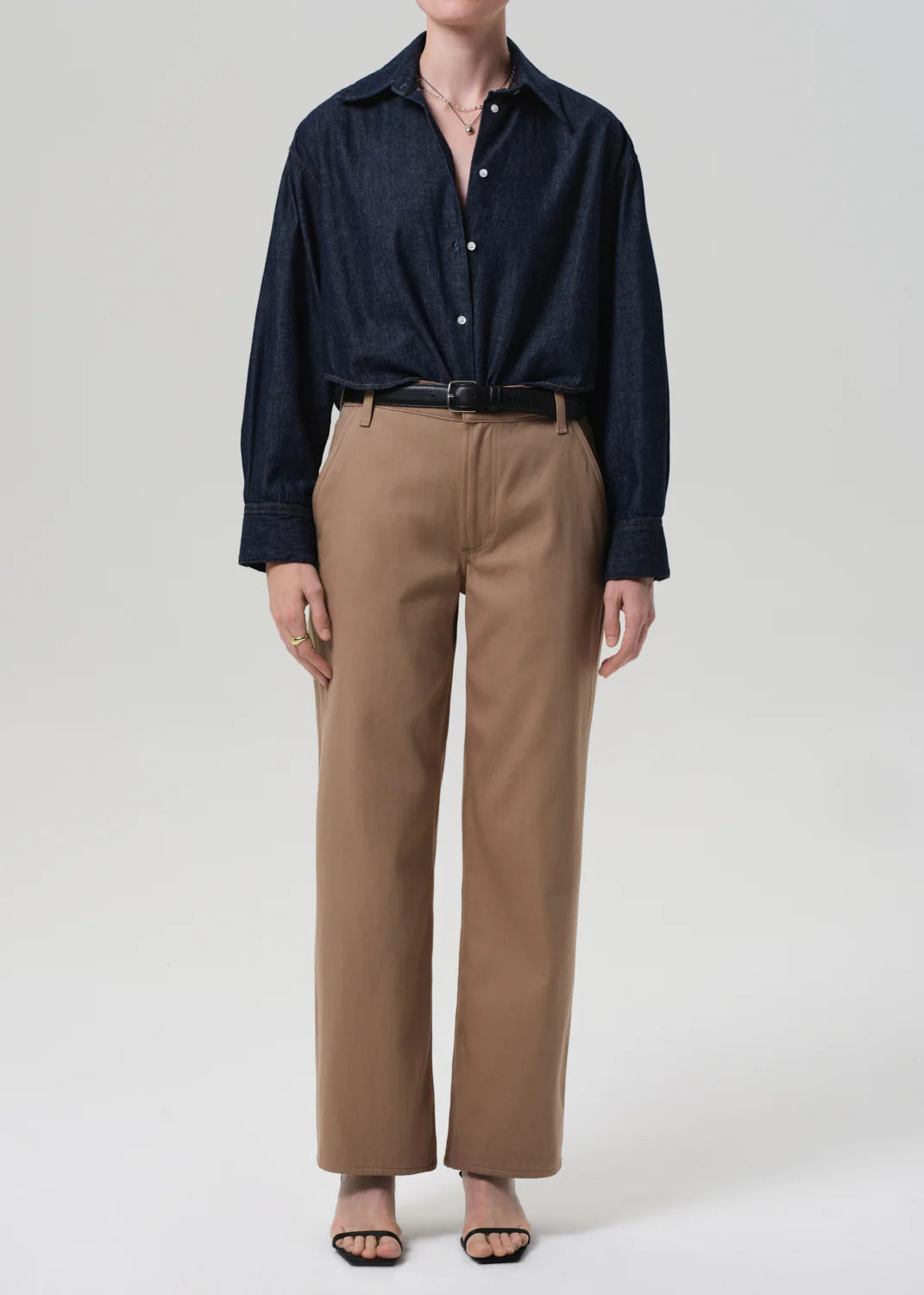 A person wears the Ana Cropped Shirt in Piave by Citizens Of Humanity/AGOLDE, tucked into high-waisted khaki trousers with a black belt, black open-toe sandals, and a delicate necklace, standing against a plain light background.