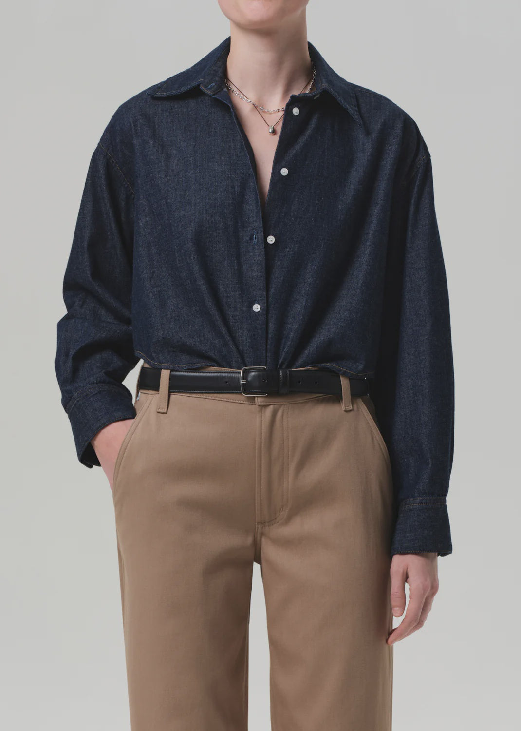 A person wears the Ana Cropped Shirt in Piave by Citizens Of Humanity/AGOLDE, a dark indigo Japanese denim button-up, tucked into high-waisted tan trousers with a black belt, one hand in their pocket and face not visible.