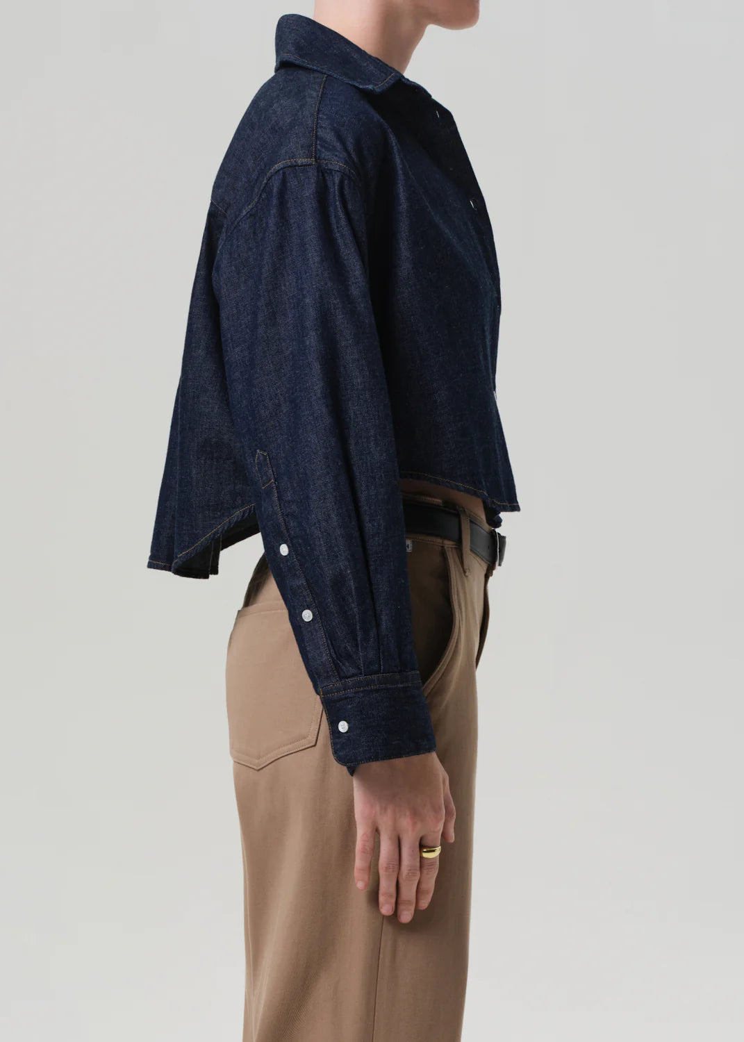 A person stands in profile wearing the Ana Cropped Shirt in Piave by Citizens Of Humanity/AGOLDE, styled with high-waisted tan trousers against a plain light gray background.