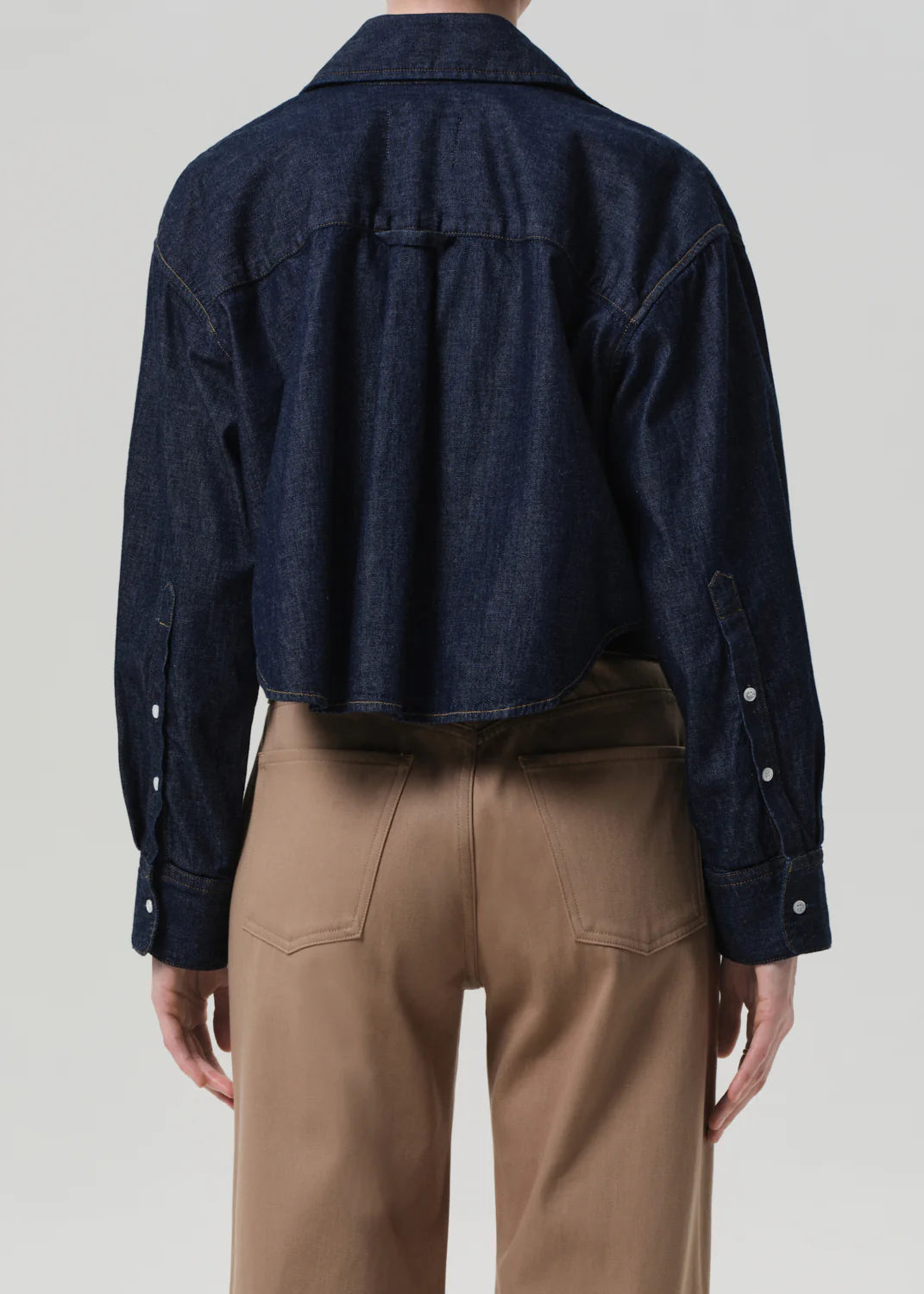A person wears the Ana Cropped Shirt in Piave by Citizens Of Humanity/AGOLDE, a dark blue Japanese denim jacket with long sleeves, paired with high-waisted tan pants, seen from the back against a plain light background.