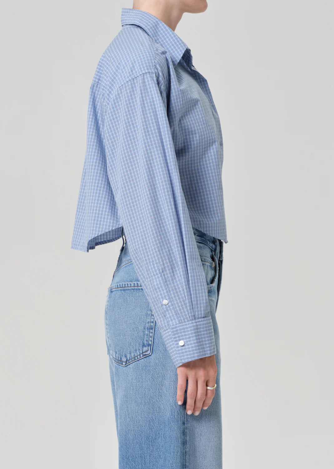 A person stands in profile wearing the ana cropped shirt in banker blue plaid by Citizens Of Humanity/AGOLDE and high-waisted light blue jeans against a plain light background.