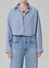 A person wears the ana cropped shirt in banker blue plaid by Citizens Of Humanity/AGOLDE, tucked into high-waisted light blue jeans, with rolled sleeves, a black pendant necklace, and rings, standing against a plain background.