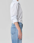 A person stands sideways in a Citizens Of Humanity/AGOLDE avni slim shirt in blue micro stripe with rolled-up sleeves, paired with high-waisted blue jeans against a plain, light background.