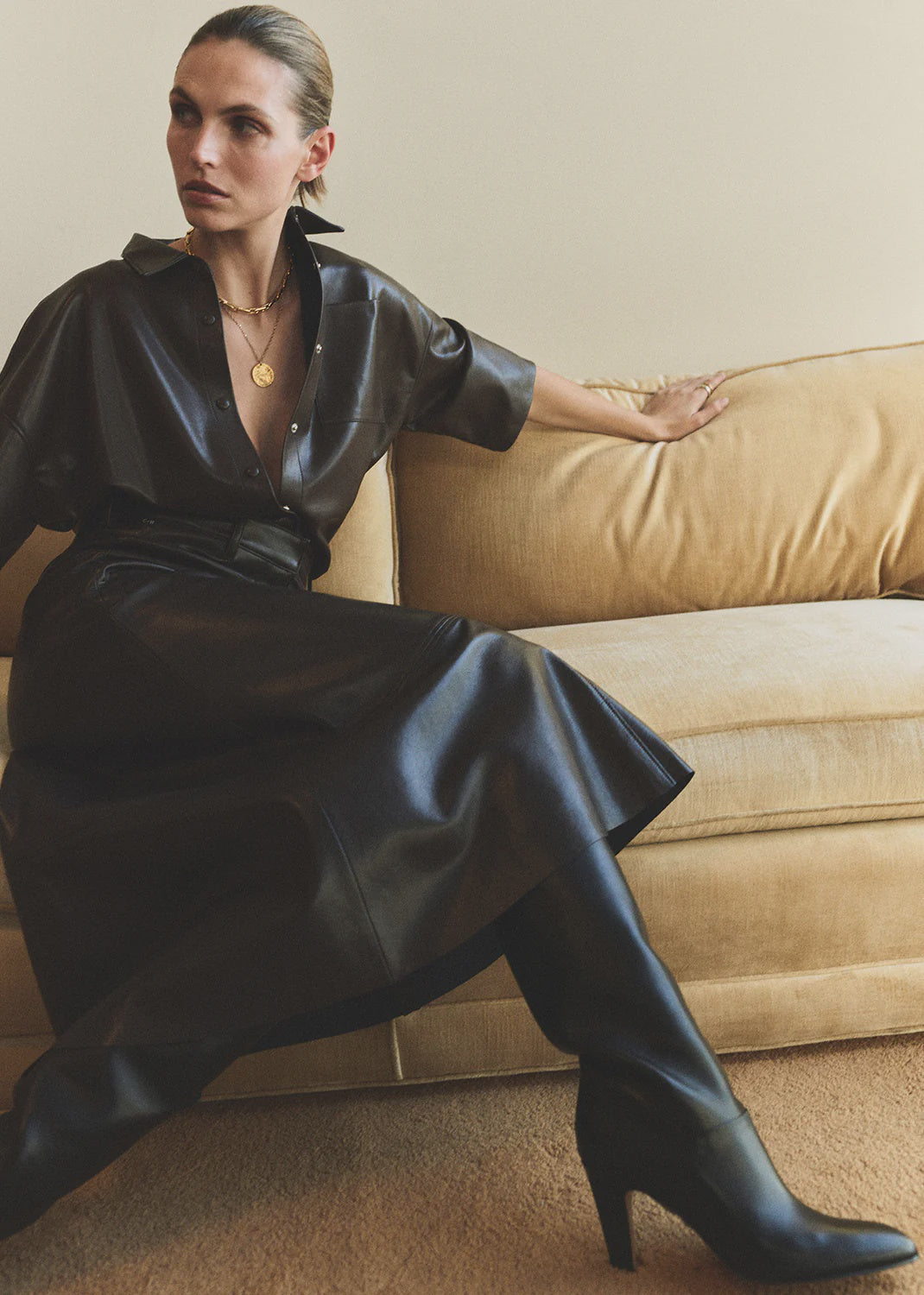 A woman sits on a beige sofa wearing the Frances Leather Cropped Cape by Citizens Of Humanity/AGOLDE, paired with black heeled boots and layered gold necklaces. She poses with one arm on the couch's back, looking to the side.