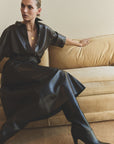 A woman sits on a beige sofa wearing the Frances Leather Cropped Cape by Citizens Of Humanity/AGOLDE, paired with black heeled boots and layered gold necklaces. She poses with one arm on the couch's back, looking to the side.