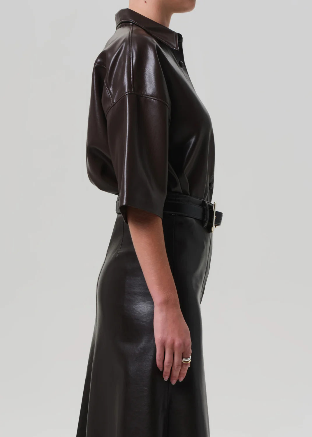 A woman in profile wears the Citizens Of Humanity/AGOLDE Frances Leather Cropped Cape in chocolate brown faux leather, featuring short sleeves and a shiny finish. Her left arm rests at her side, displaying a thin ring. The backdrop is light gray.