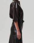 A woman in profile wears the Citizens Of Humanity/AGOLDE Frances Leather Cropped Cape in chocolate brown faux leather, featuring short sleeves and a shiny finish. Her left arm rests at her side, displaying a thin ring. The backdrop is light gray.