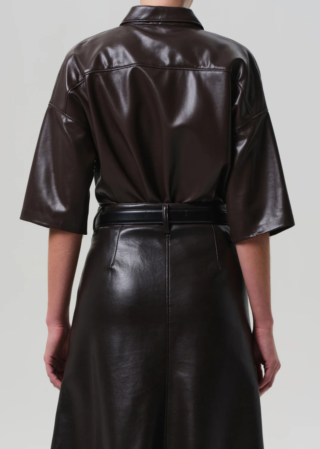 A person is seen from behind wearing the Frances Leather Cropped Cape by Citizens Of Humanity/AGOLDE, featuring a chocolate brown glossy faux leather, elbow-length sleeves, belted waist, and set against a plain light gray background.