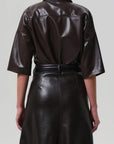 A person is seen from behind wearing the Frances Leather Cropped Cape by Citizens Of Humanity/AGOLDE, featuring a chocolate brown glossy faux leather, elbow-length sleeves, belted waist, and set against a plain light gray background.