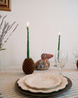 A striped tablecloth holds a white plate, bowl, and glass. Two lit Faire Asparagus Beeswax Candles in pinecone holders flank a ceramic bird centerpiece, while a vase with lavender stems sits to the left.
