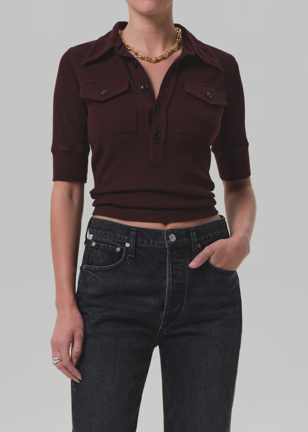 Wearing the Amina Polo Shirt by Citizens Of Humanity/AGOLDE—a dark burgundy, ribbed cotton short-sleeve collared top with buttons—paired with high-waisted black jeans and a gold chain necklace against a plain background.