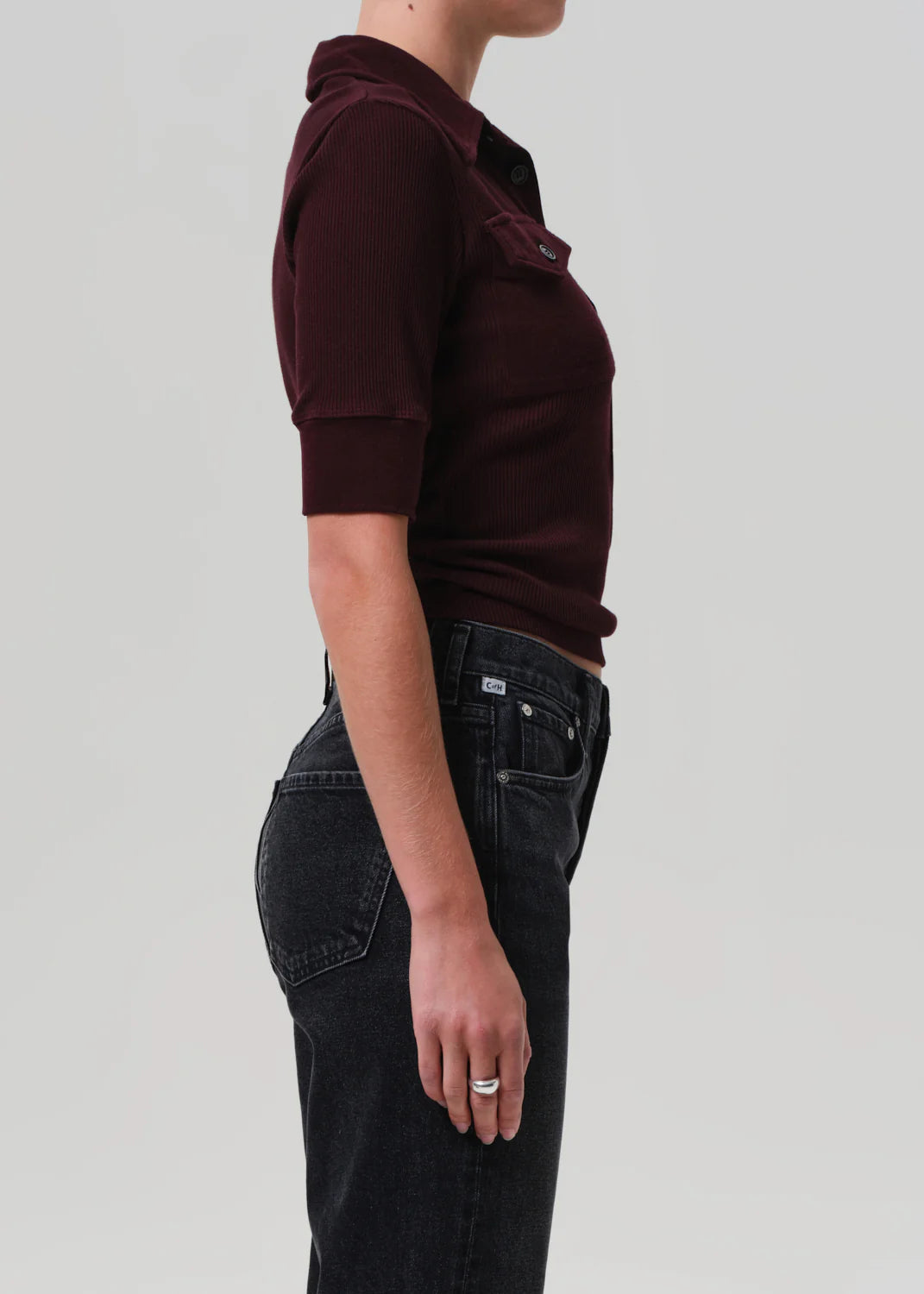 A person stands in profile against a plain background, wearing the Amina Polo Shirt by Citizens Of Humanity/AGOLDE— a ribbed cotton, short-sleeve burgundy top with collar and pocket—paired with high-waisted black jeans. Their left arm is relaxed by their side.
