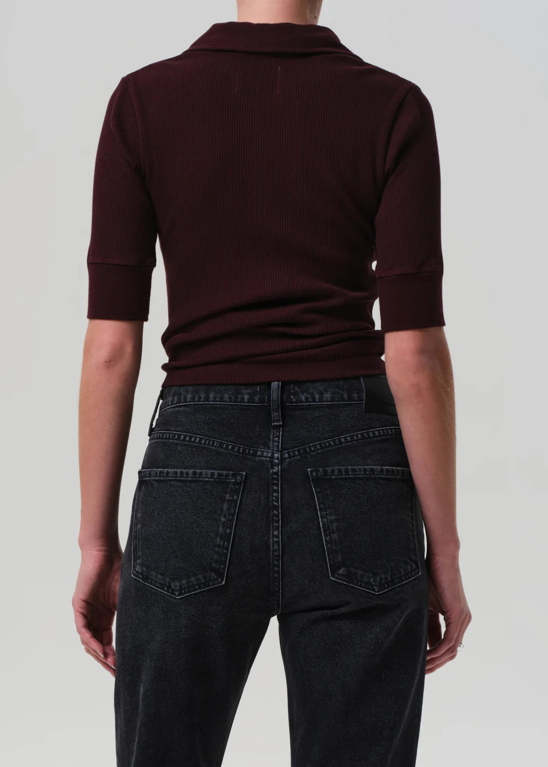 A person stands with their back to the camera, wearing the Amina Polo Shirt by Citizens Of Humanity/AGOLDE in dark burgundy ribbed cotton and high-waisted black denim jeans against a plain, light background.