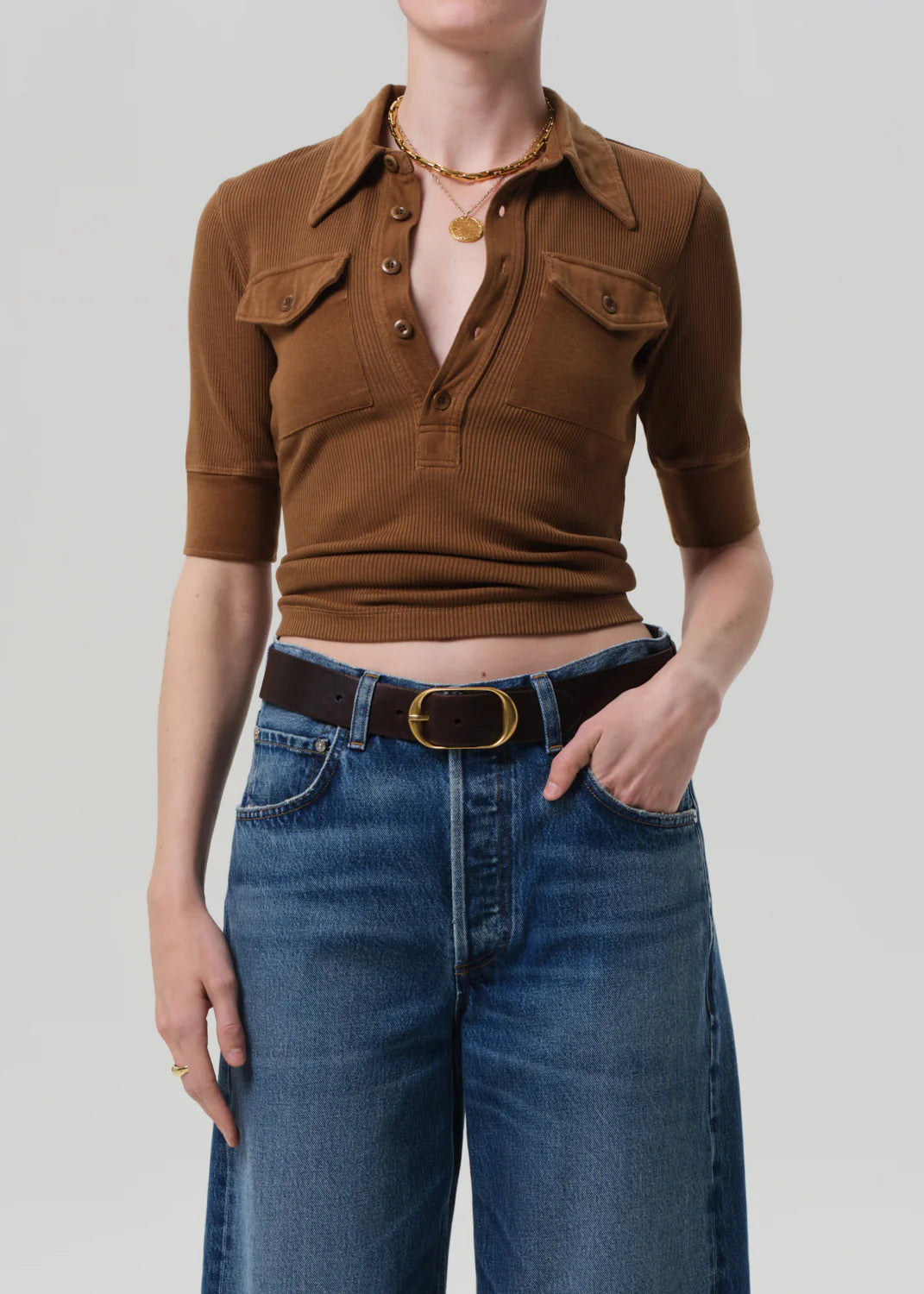 A person wears the Citizens Of Humanity/AGOLDE Amina Polo Shirt—ribbed cotton, brown with front patch pockets—tucked into high-waisted blue jeans with a black belt and gold buckle, finished with layered gold necklaces and a ring.