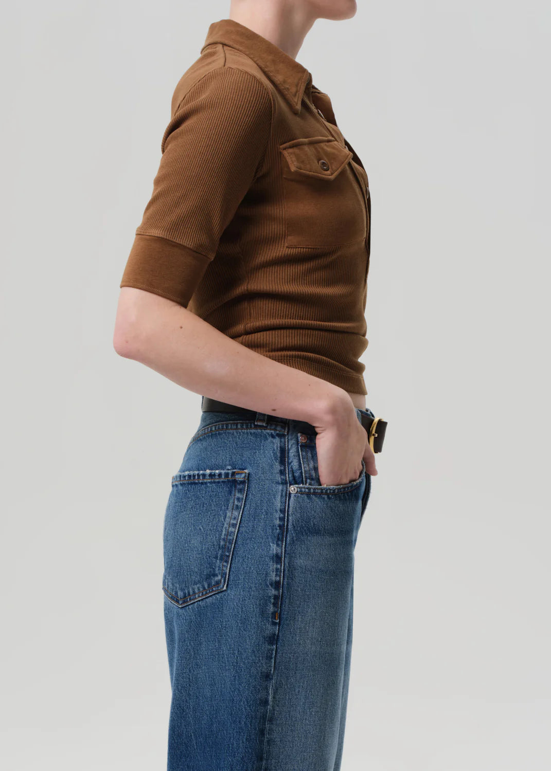 A person in the Citizens Of Humanity/AGOLDE Amina Polo Shirt and blue jeans with front patch pockets stands in profile with one hand in their pocket against a plain light gray background.