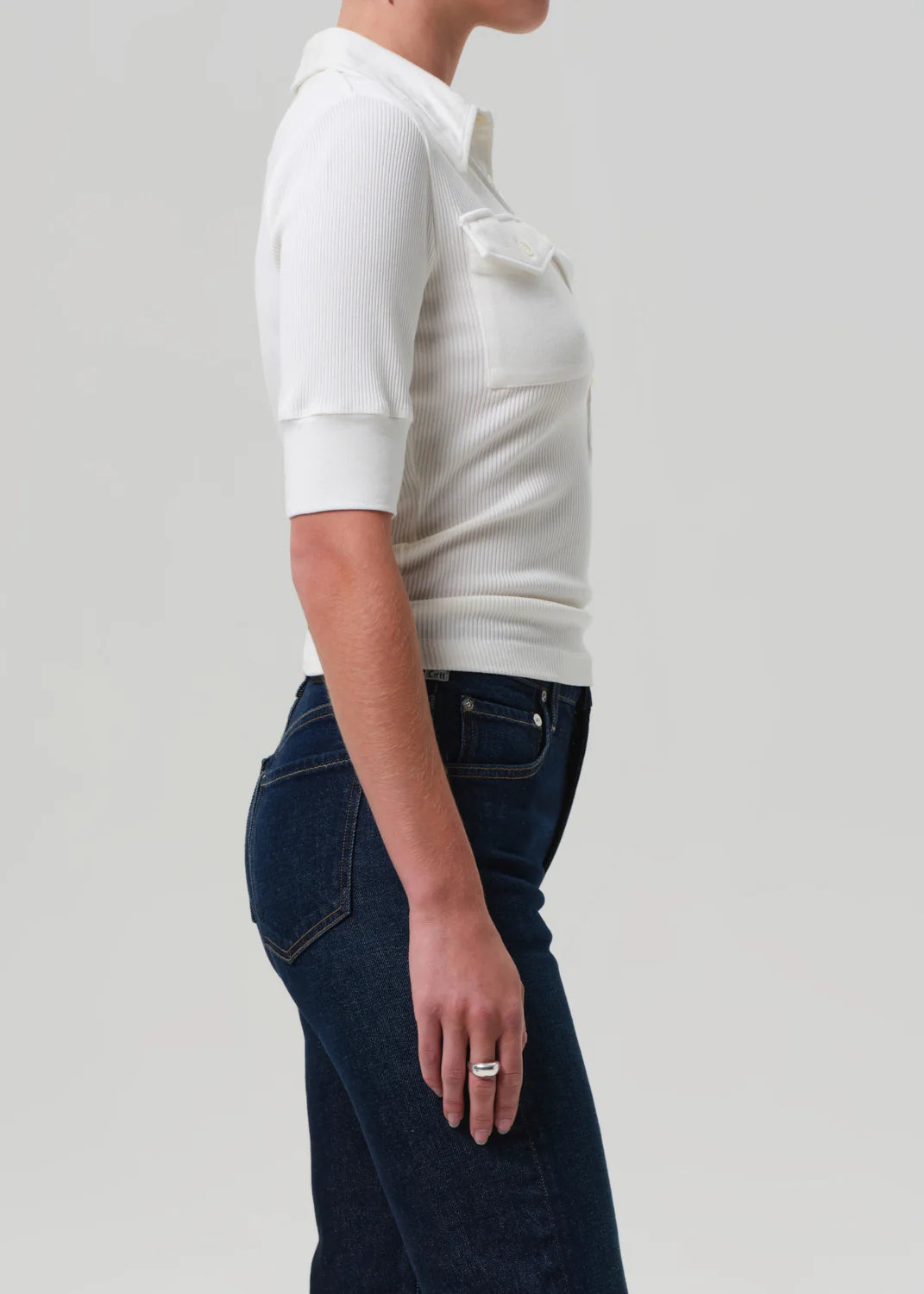 A person in profile wears the Amina Polo Shirt by Citizens Of Humanity/AGOLDE, a white ribbed cotton, short-sleeve top with a collar and front pocket detail, paired with dark blue jeans against a plain light background.