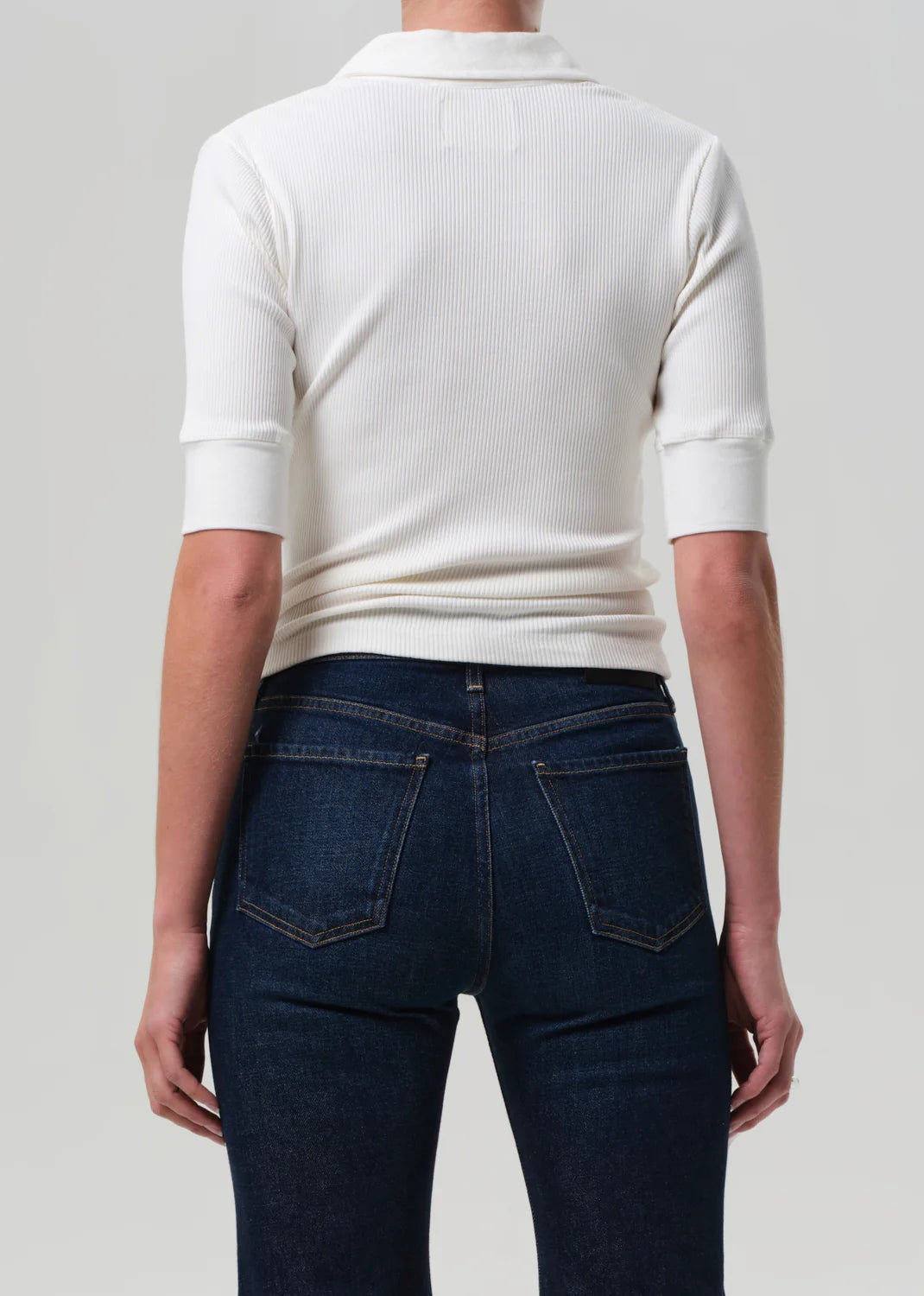 A person is shown from the back, wearing the Amina Polo Shirt by Citizens Of Humanity/AGOLDE in white, styled with dark blue jeans with front pockets. The background is plain and light gray.