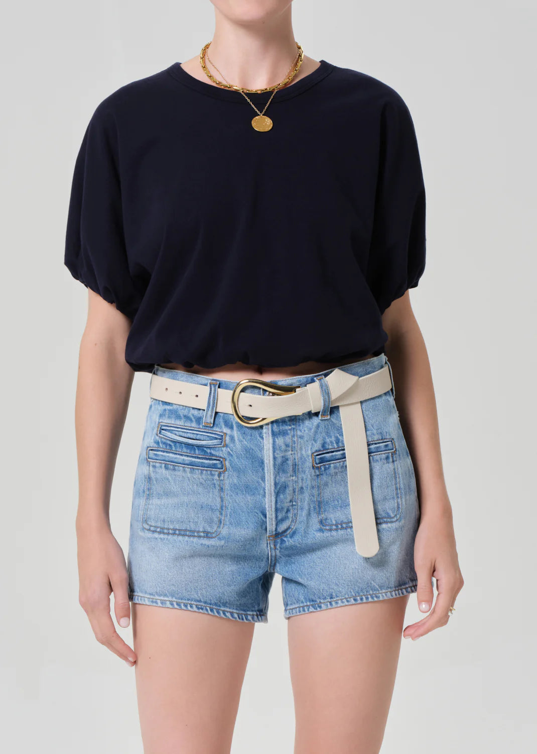 Wearing the Citizens Of Humanity/AGOLDE gia bubble tee tucked into high-waisted light blue denim shorts with front pockets, styled with a wide white belt, gold necklace, and ring against a plain light background.