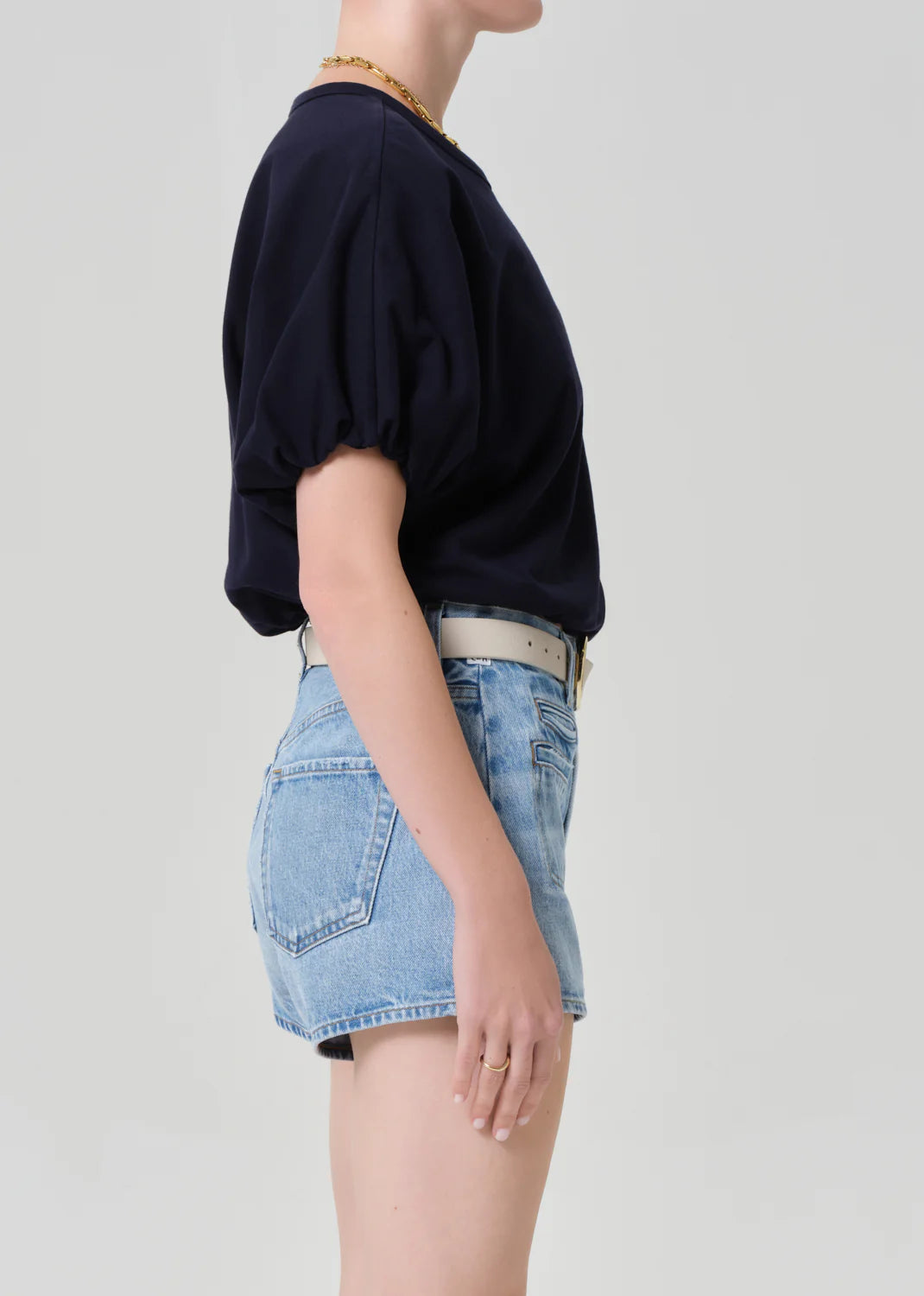 A person stands in profile against a plain background, wearing the Citizens Of Humanity/AGOLDE gia bubble tee in navy, light blue high-waisted denim shorts, a white belt, and a gold necklace.
