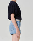 A person stands in profile against a plain background, wearing the Citizens Of Humanity/AGOLDE gia bubble tee in navy, light blue high-waisted denim shorts, a white belt, and a gold necklace.