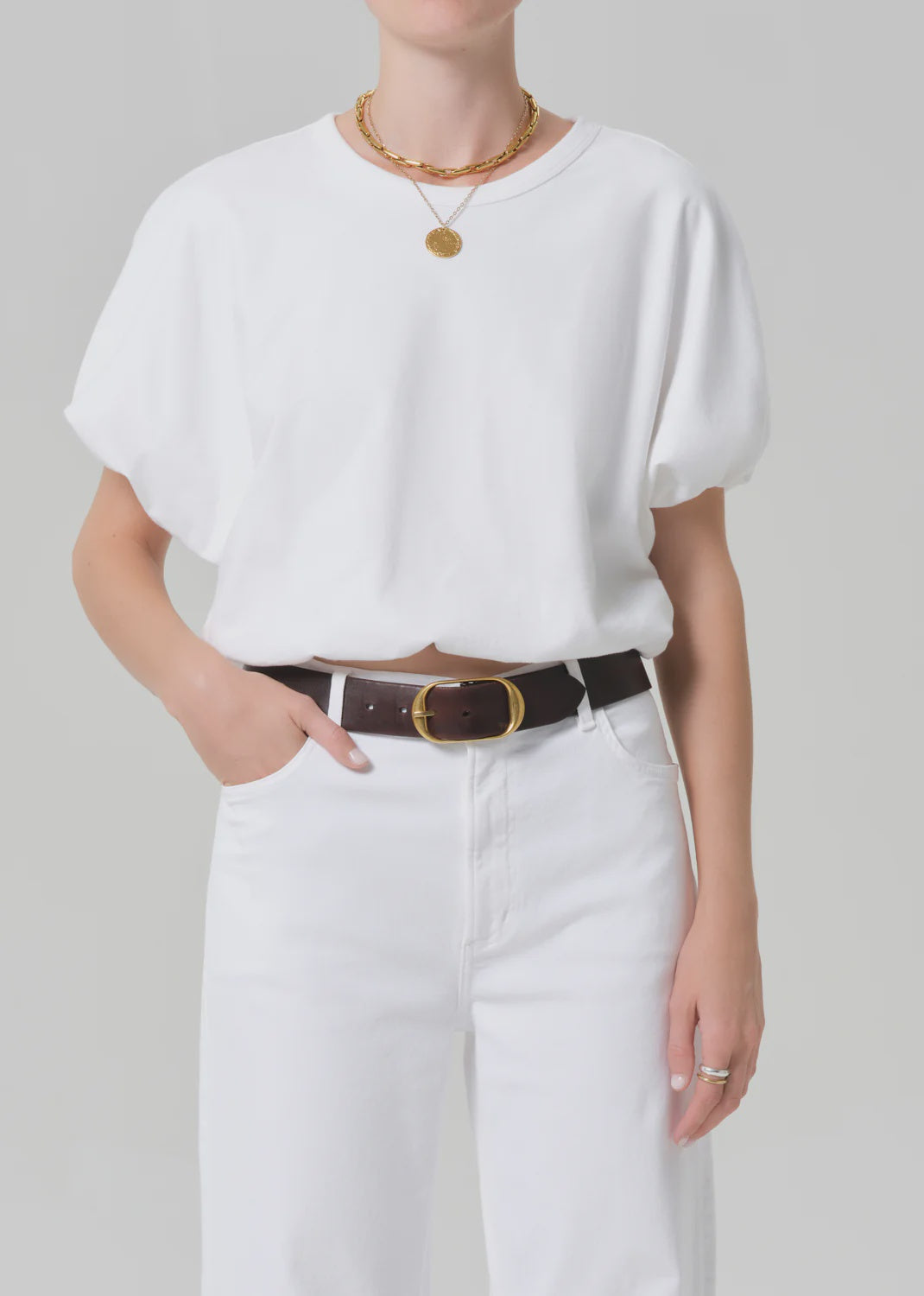 Model wears the gia bubble tee by Citizens Of Humanity/AGOLDE—white cotton, tucked into white jeans with a dark brown belt, gold necklaces, and a ring. Upper body shown against a plain light gray background.