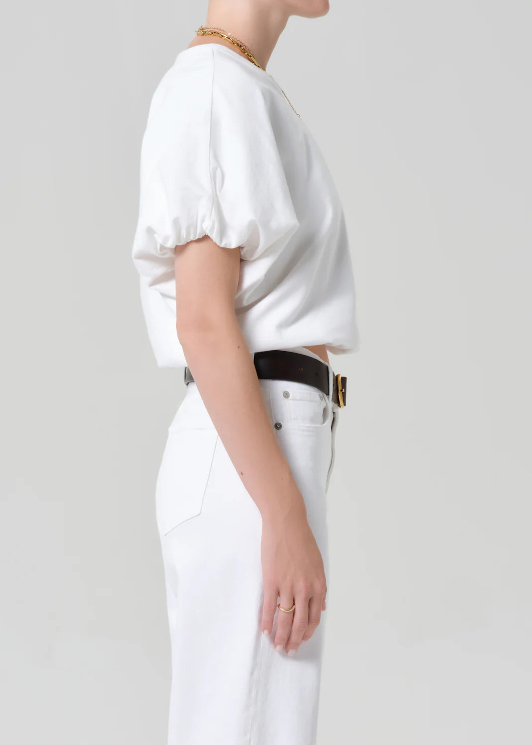 A person in profile wears the Citizens Of Humanity/AGOLDE gia bubble tee with puff sleeves, white high-waisted pants, a black belt with gold buckle, a gold chain necklace, and a ring against a plain light grey background.