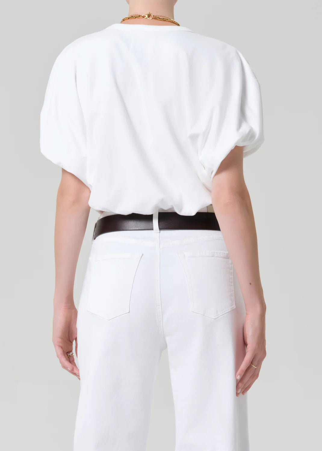 A person faces away, wearing the Citizens Of Humanity/AGOLDE gia bubble tee—a loose white jersey cotton short-sleeve top—tucked into high-waisted white jeans with a black belt. The background is plain and light-colored.
