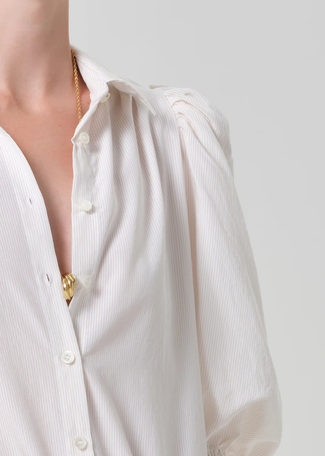 Wearing the Anya Puff Sleeve Shirt by Citizens Of Humanity/AGOLDE, a loose white blouse with thin stripes and a partially unbuttoned top that reveals a gold necklace, stands out against a plain light-colored background.