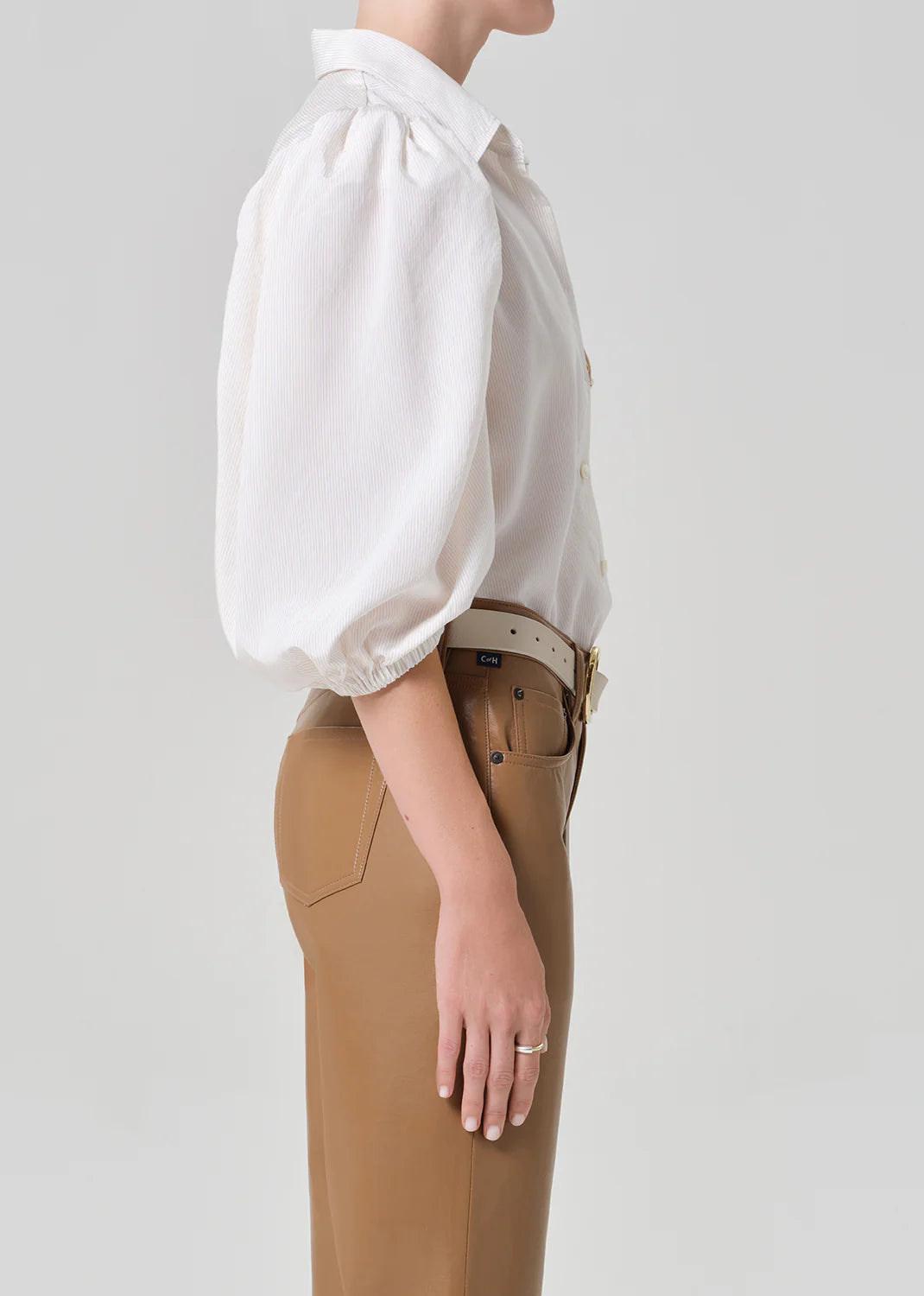 A person stands in profile wearing the Citizens Of Humanity/AGOLDE Anya Puff Sleeve Shirt tucked into high-waisted tan trousers, accessorized with a white belt and a ring, set against a plain light background.
