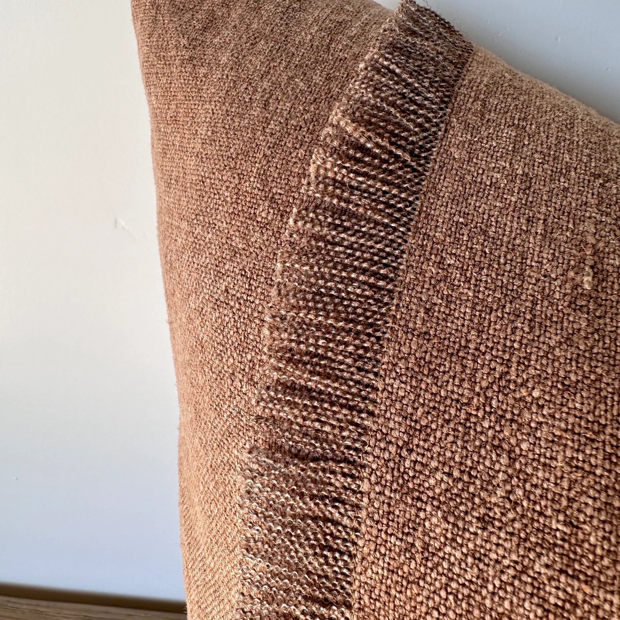 A close-up of the Fringe Accent Pillow Havane 22x22, featuring a rust brown hue and vertical stripe, propped against a white wall. Its linen fabric showcases a soft woven texture and detailed stitching.