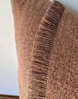 A close-up of the Fringe Accent Pillow Havane 22x22, featuring a rust brown hue and vertical stripe, propped against a white wall. Its linen fabric showcases a soft woven texture and detailed stitching.